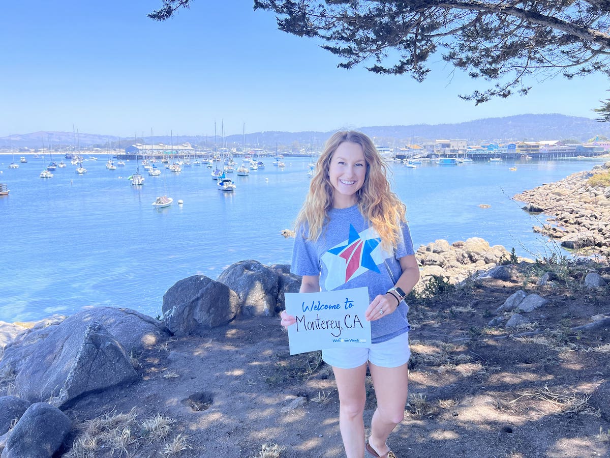 Blue Star Volunteer Misty stands at an oceanfront with a Blue Star Families T-Shirt and a sign that reads Welcome to Monterey, CA