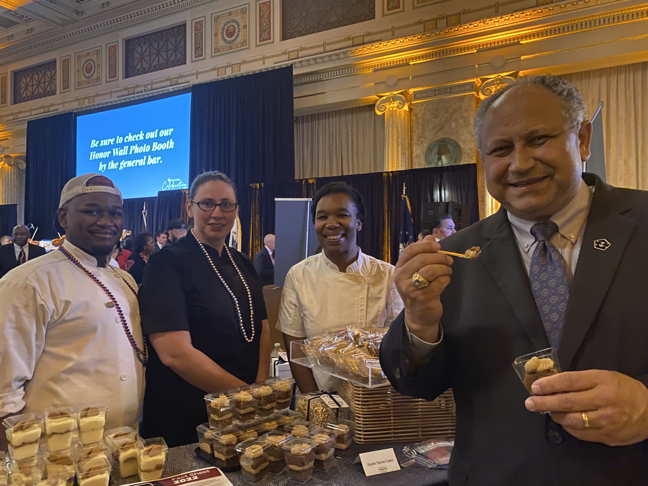 Secretary of the Navy, Carlos Del Toro, poses in front of the Dog Tag Bakery table, sampling a dessert.