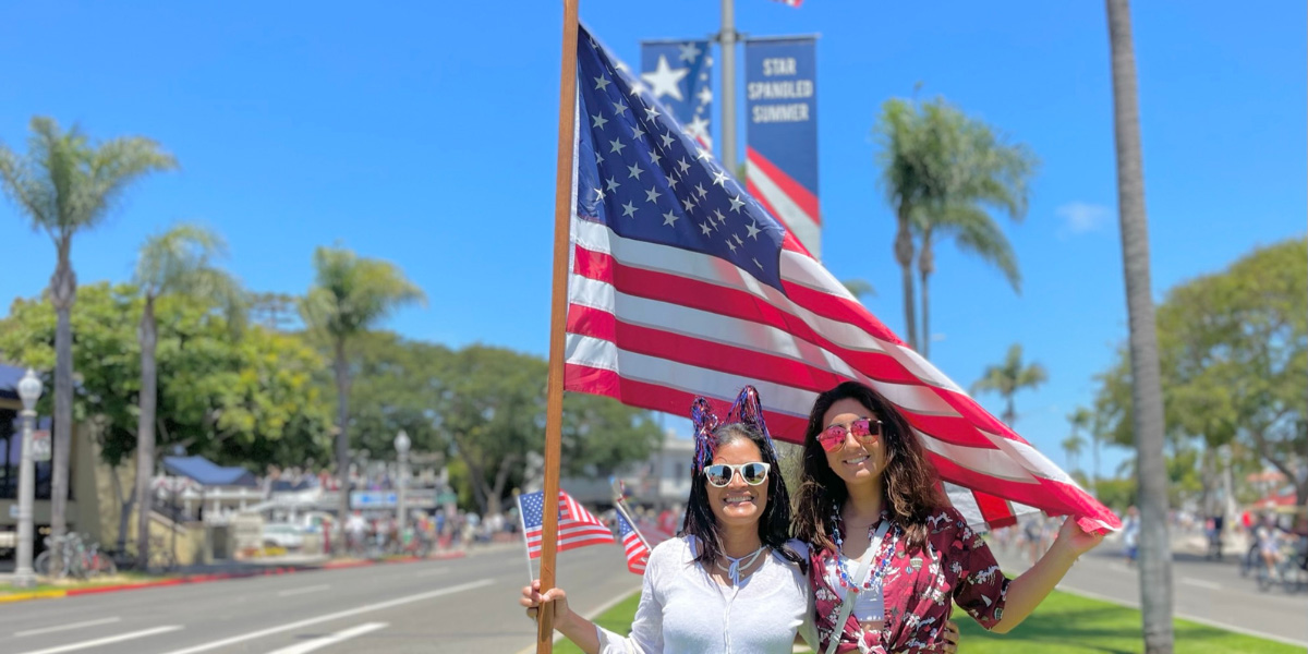 Two women hold an American flag over their heads while standing on a grassy street median. 