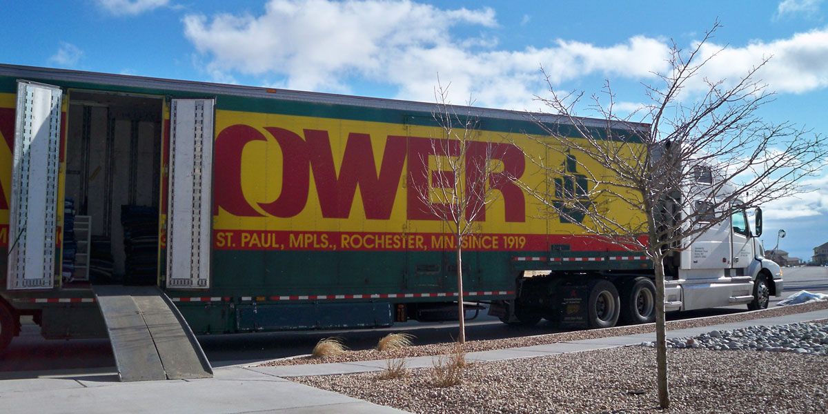 A green moving truck is parked outside with its hind doors open and a ramp leading into the back of the truck. The word “Mayflower” is written in red on a yellow background, with a green sailing ship to the right side of the back of the truck.