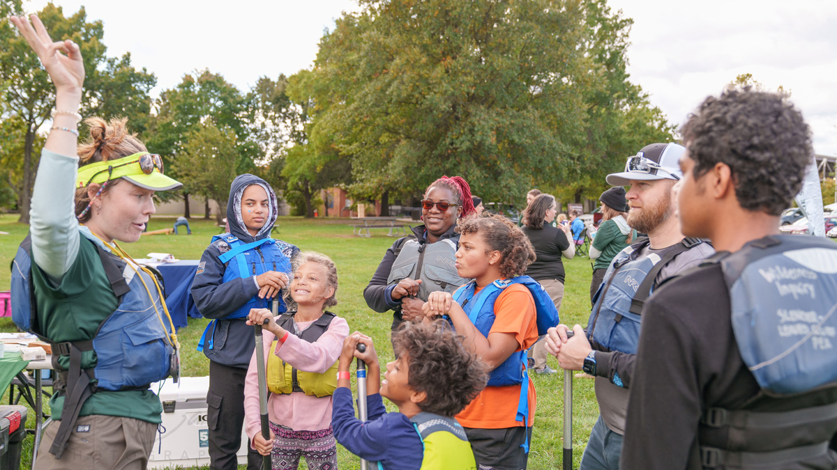 A group of adults and children gathered outdoors in a park wearing life jackets. One adult, standing on the left, is raising her hand and explaining something to the group.