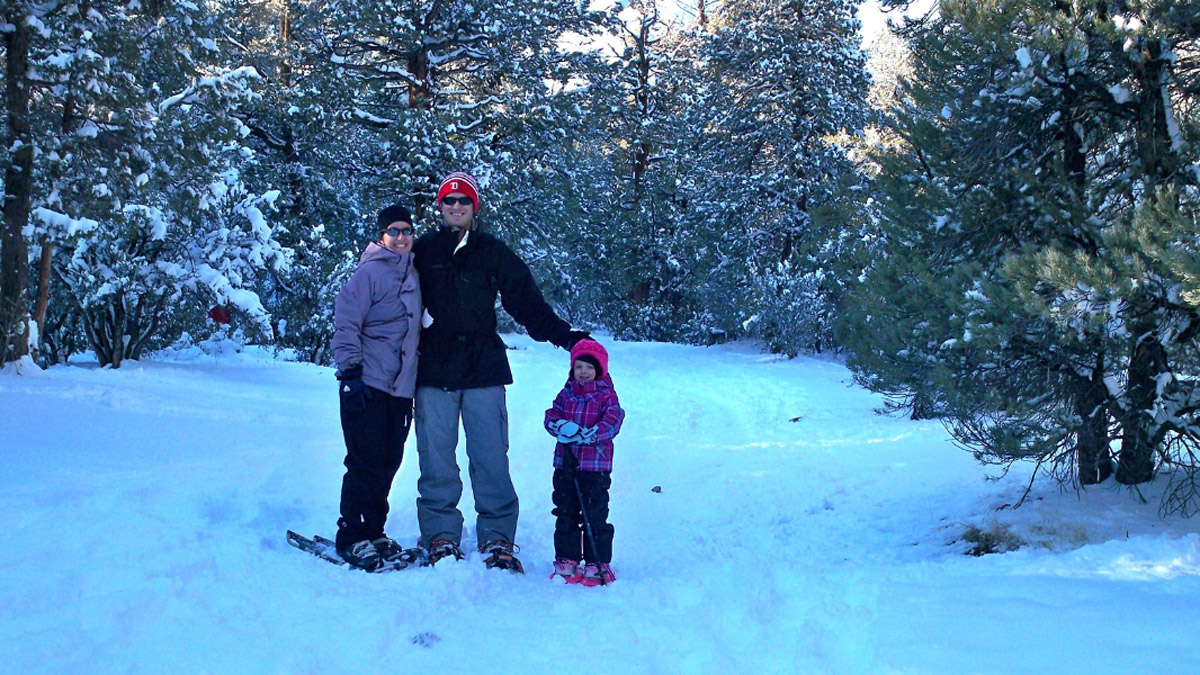 Three people wearing winter weather clothing and snowshoes. Person on the right is a small child. In the background are evergreen trees and the ground is covered in snow. 