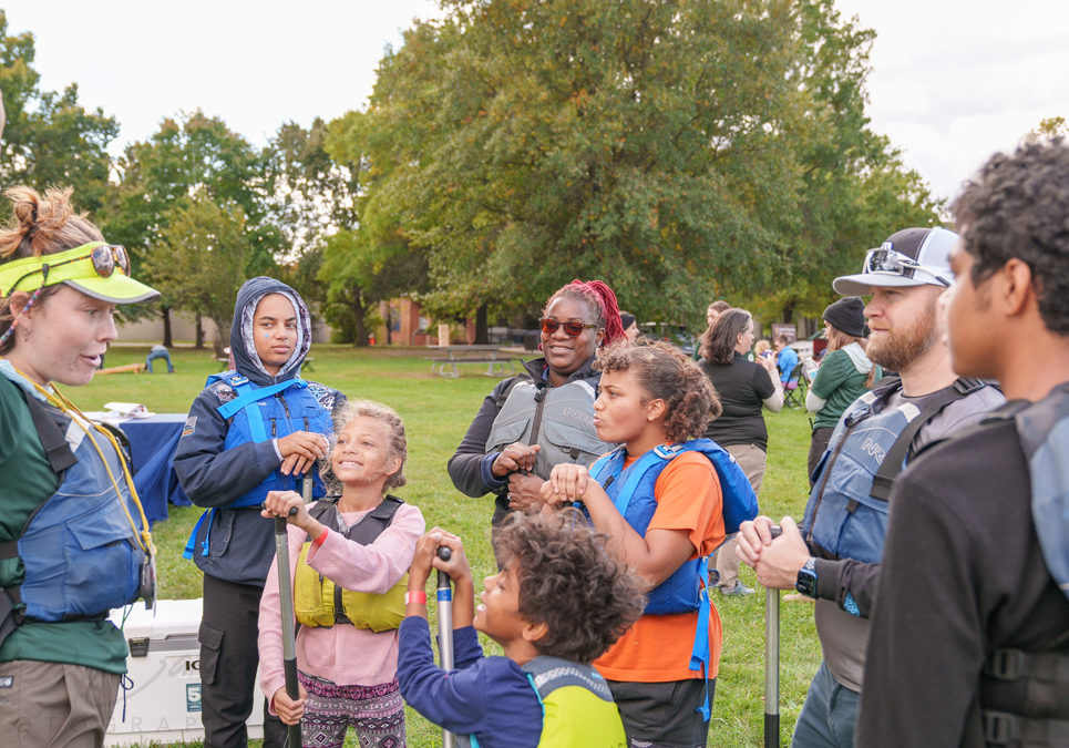 A group of adults and children gathered outdoors in a park wearing life jackets. One adult, standing on the left, is raising her hand and explaining something to the group.