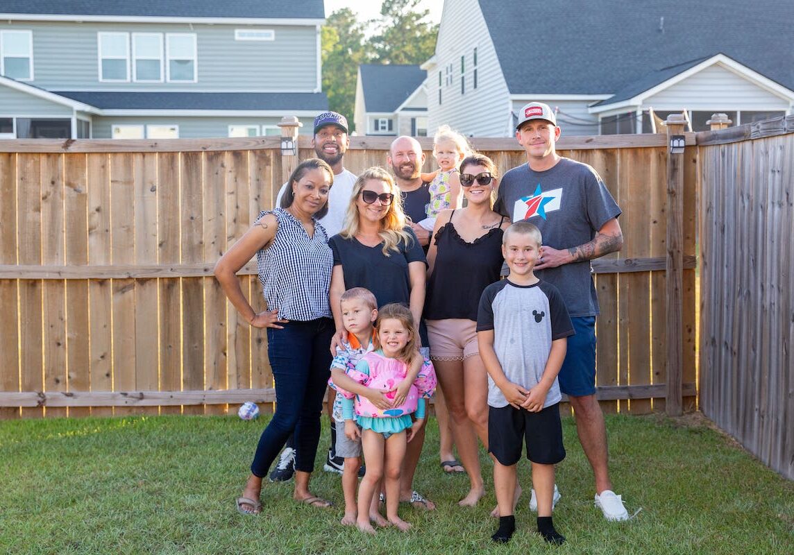 Adults and children stand, posing together in front of a tall fence in a backyard.