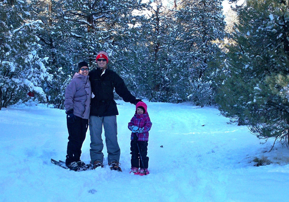 Three people wearing winter weather clothing and snowshoes. Person on the right is a small child. In the background are evergreen trees and the ground is covered in snow.